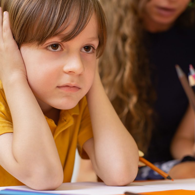 Close up of a cute little boy looking upset at school. Sad young boy sitting in classroom. Little boy looking bored during school lesson. Boredom, tiredness concept